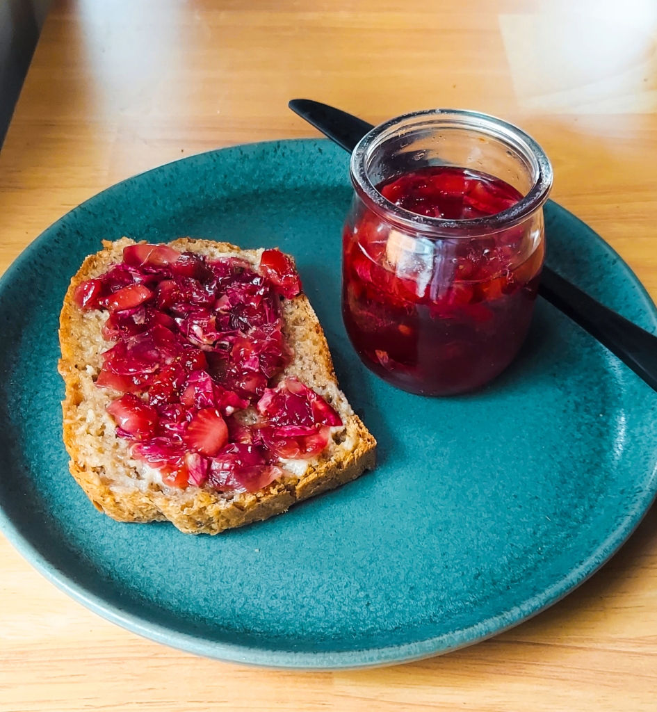 A piece of bread covered in strawberry and rose petal jam, sitting on a blue-green plate beside a jar of said jam and a butter knife. 