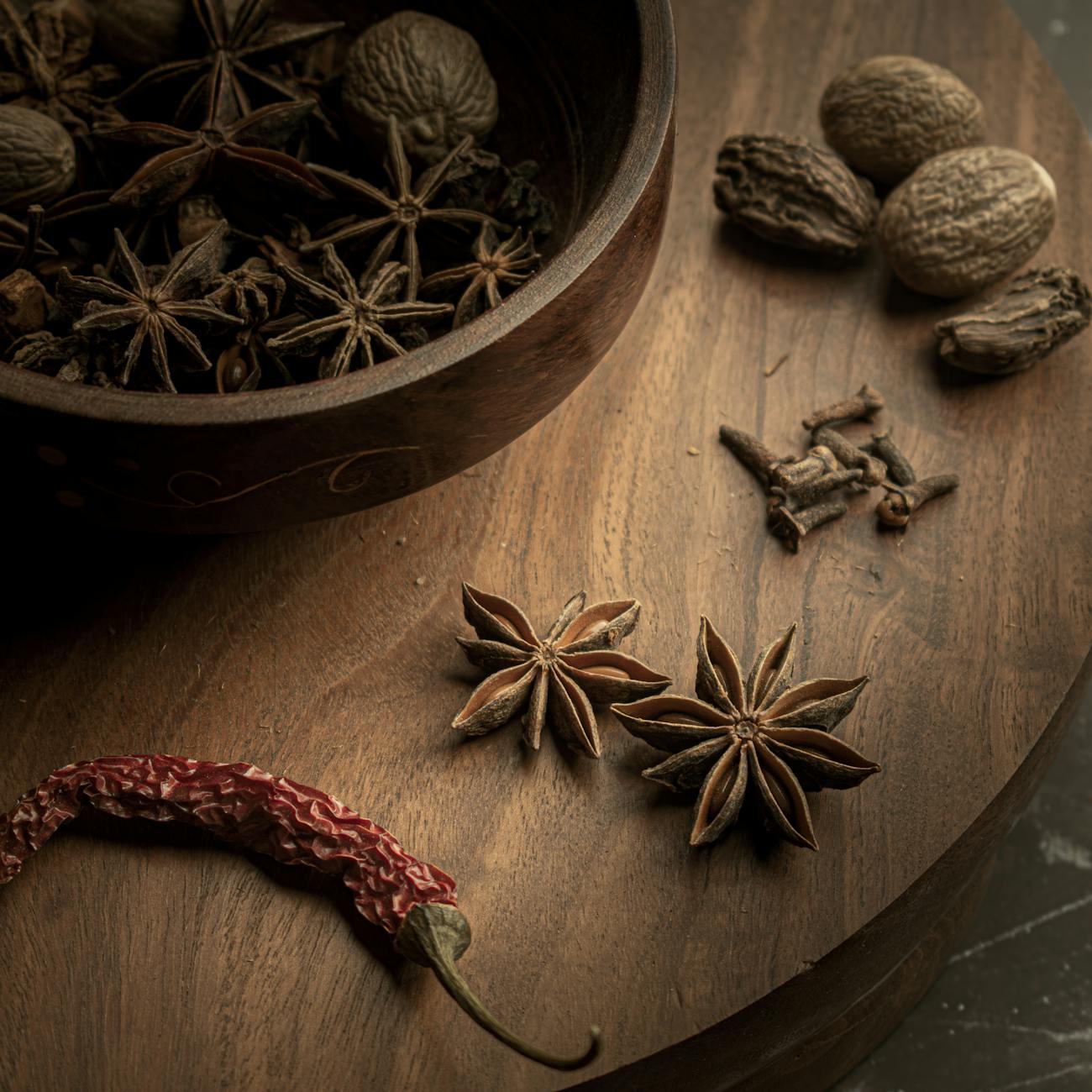 A dried chili pepper, whole star anise, clove buds, and whole nutmegs on a wooden table. 